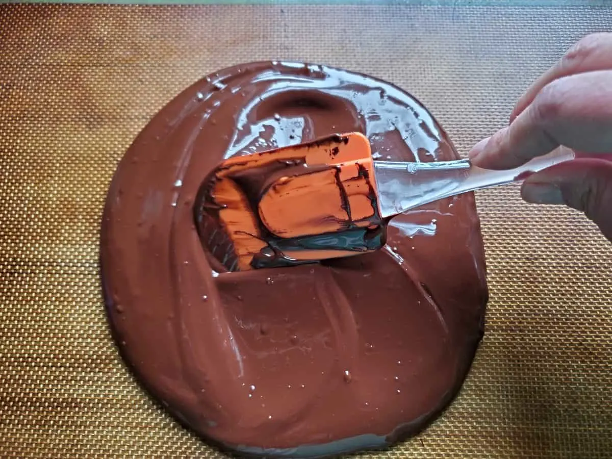 Orange spatula spreading melted chocolate on a silpat.