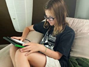 Girl in a black t-shirt sitting on a stuffed chair playing on a Kindle.