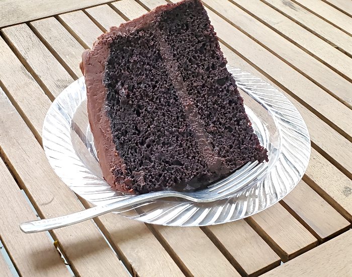 Slice of chocolate cake on a plate with a fork sitting on a wooden slat table.