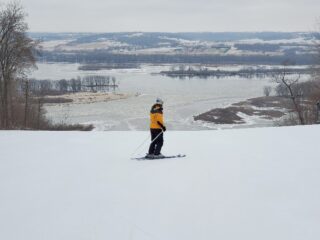 Lone woman at the top of a hill on skis overlooking a frozen river.