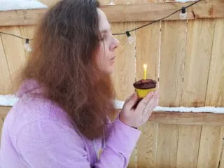 Woman blowing out a candle on a chocolate frosted cupcake by a wooden fence.