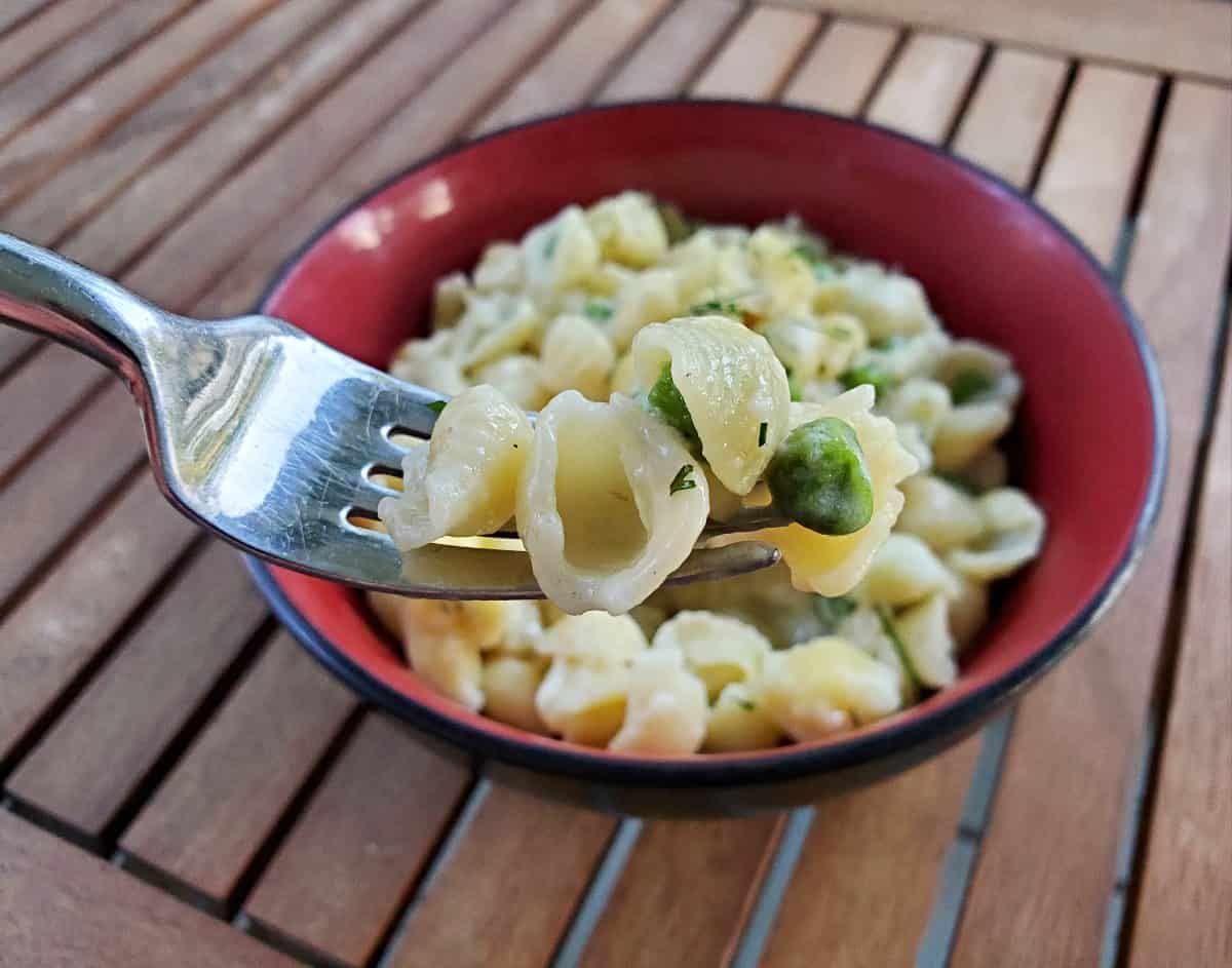 Fork of mini shell pasta with peas and dill over a bowl containing more pasta.
