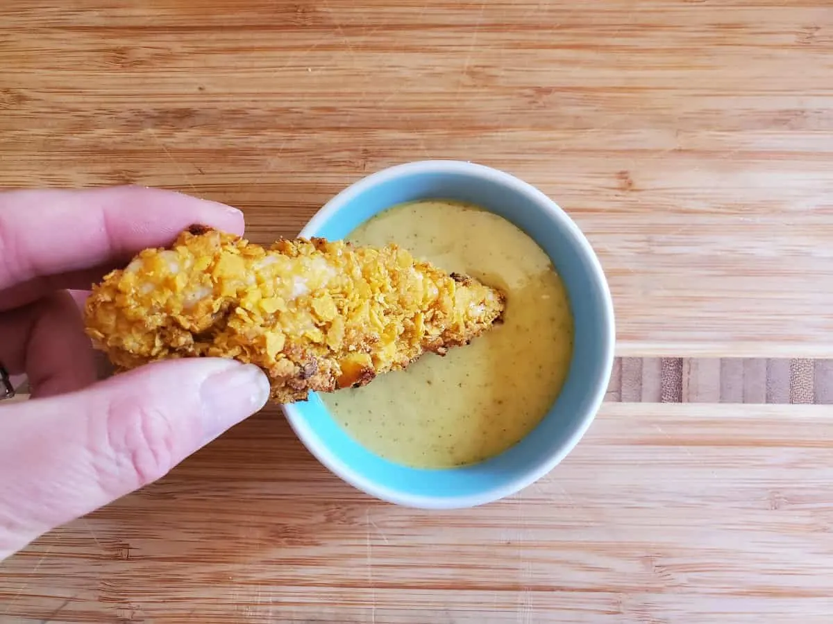 Image shows Hand dipping a corn flake crusted chicken tender into a bowl of sauce on a wooden table.