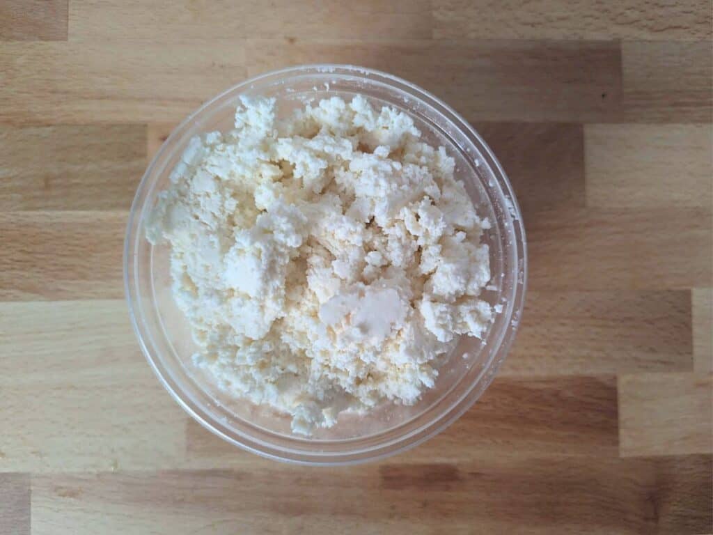 Image shows an overhead shot of ricotta in a deli container on a wooden board.
