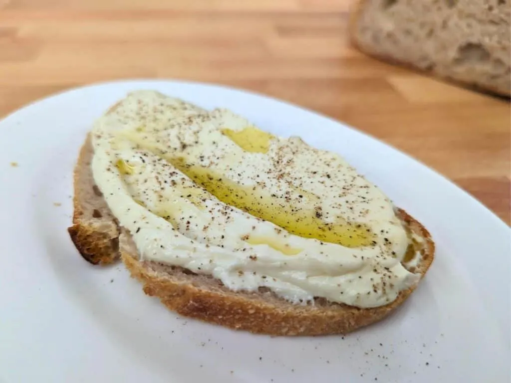 Image shows a Slice of whipped ricotta toast on a white plate on a wooden table.