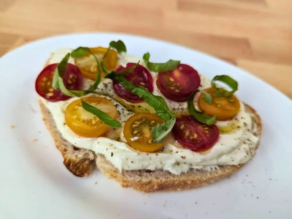 Image shows a slice of Tomato and basil on whipped ricotta toast on a white plate.