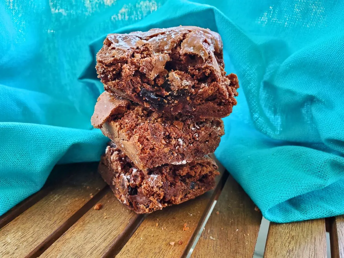 Image shows Three cherry brownies stacked on a wooden table with a teal background.