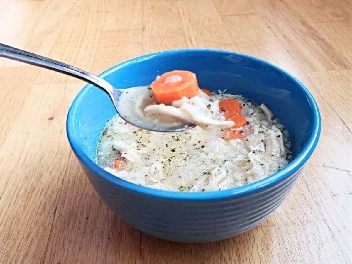 Image shows Chicken and rice soup on a spoon with the full blue bowl behind it.