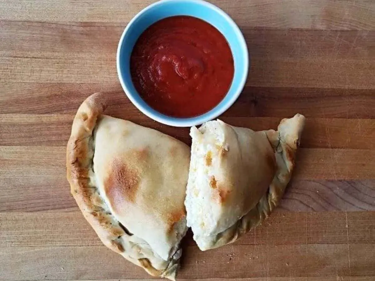 Image shows an overhead shot of a Calzone cut in half on a wooden board with a small bowl of marinara above it.