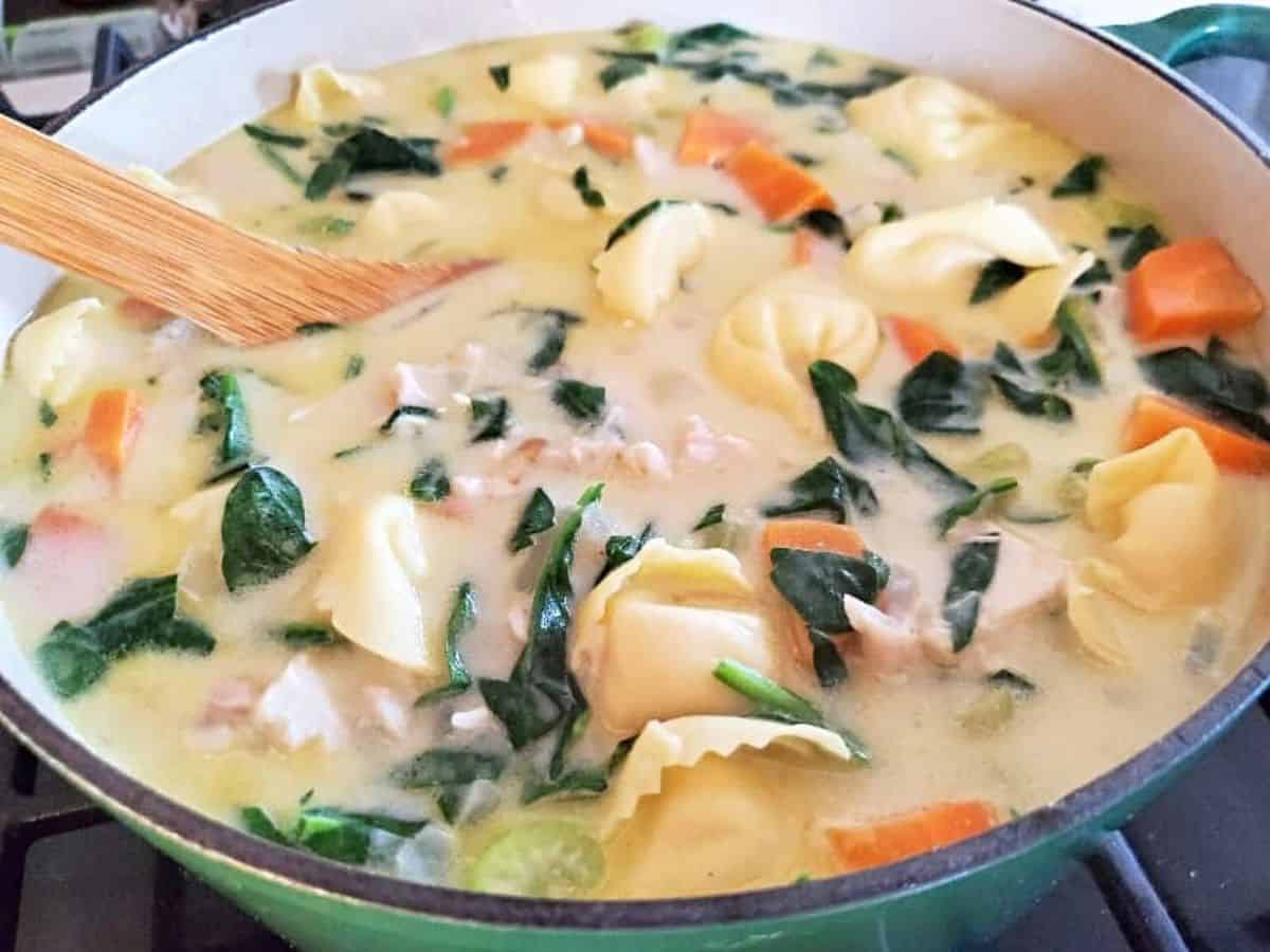 Image shows Chicken tortellini soup in a ceramic enameled pot with a wooden spoon stirring it.