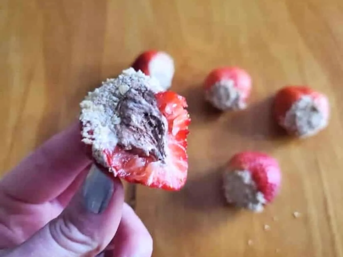 Image shows a hand holding a s'mores stuffed strawberry half with the filling exposed and more behind it.