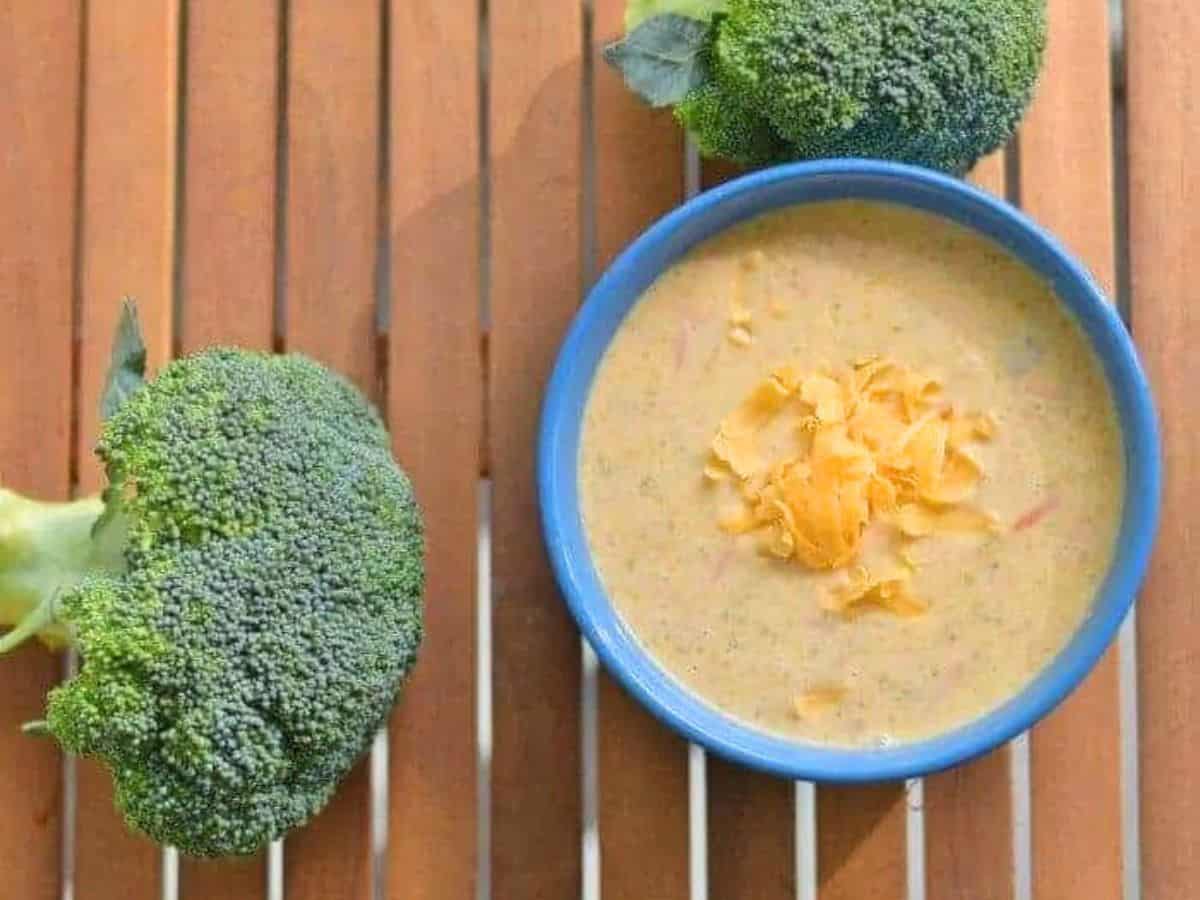 Image shows A bowl of broccoli cheddar soup with broccoli around it.