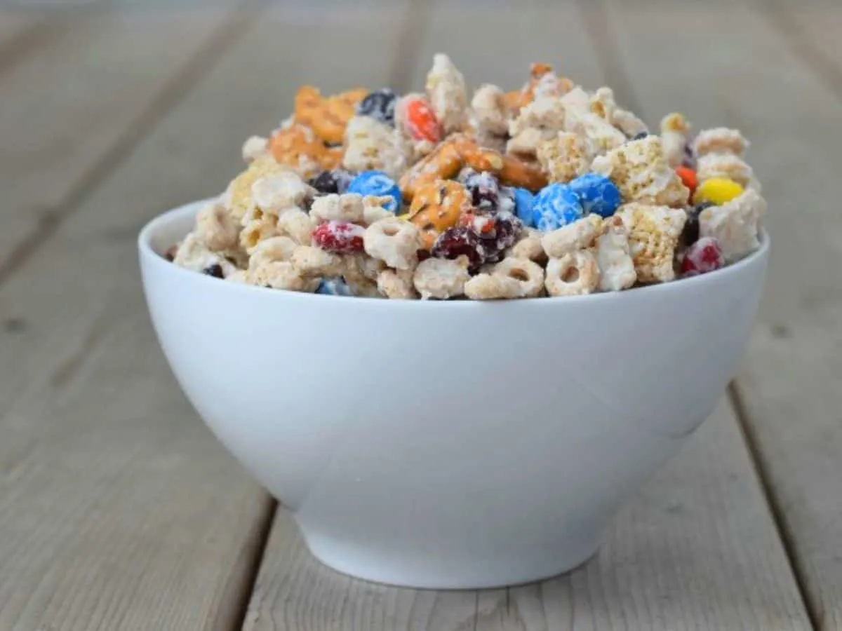 Image shows white chocolate party mix piled into a white bowl on a wooden table.