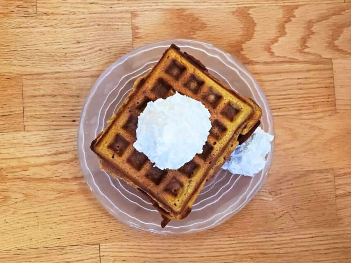 Image shows an overhead shot of Pumpkin Waffles with whipped cream on a glass plate.