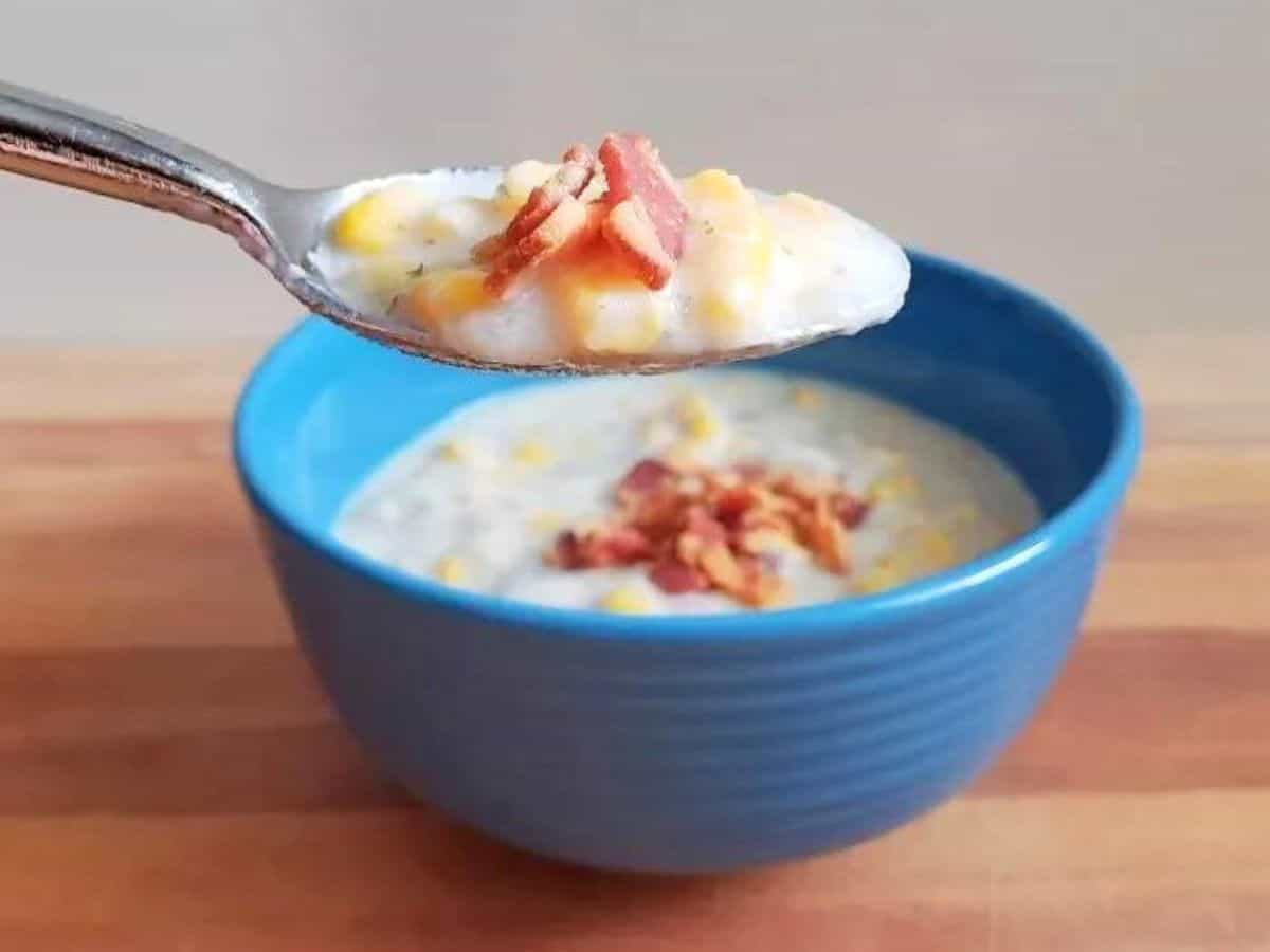 Spoon holding potato and corn chowder with bacon with the full bowl of soup behind it.