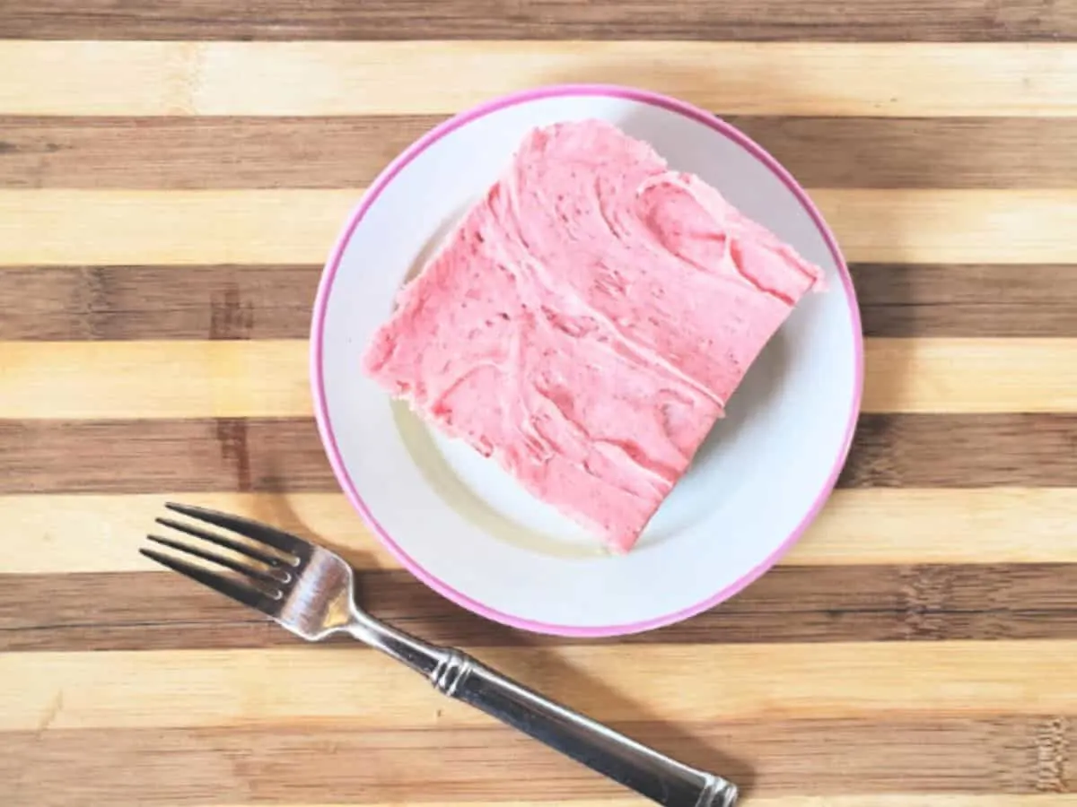 Overhead shot of a slice of cake with strawberry buttercream frosting with a fork sitting next to the plate.