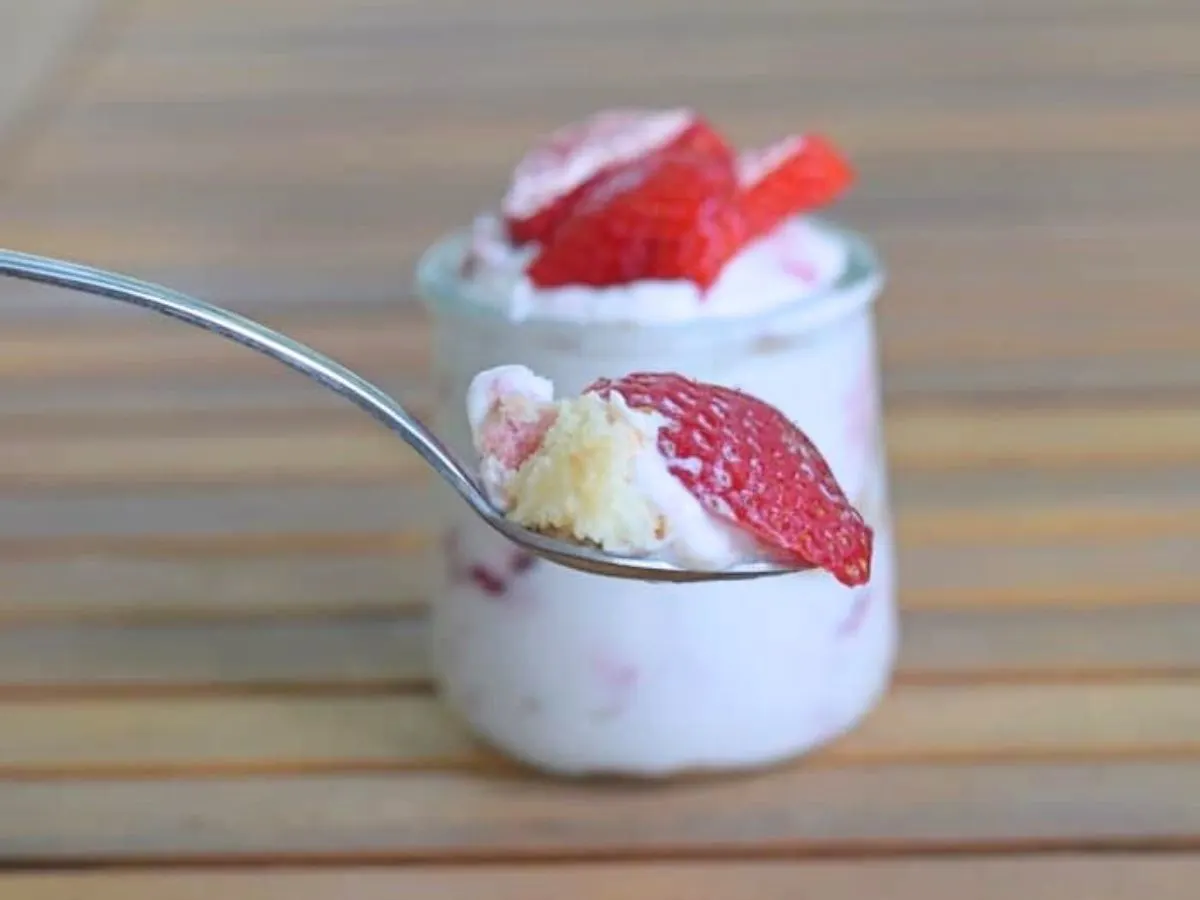 Image shows a spoon holding a scoop of the strawberry lemonade icebox cake with a small jar of the dessert behind it.