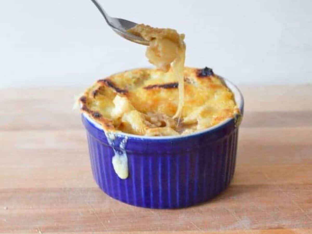 Image shows a Spoon pulling cheese from a bowl of French onion soup in a blue ramekin.