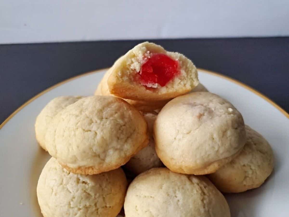 Image shows a hand holding a Cherry Surprise Cookie with more on a white plate behind it.