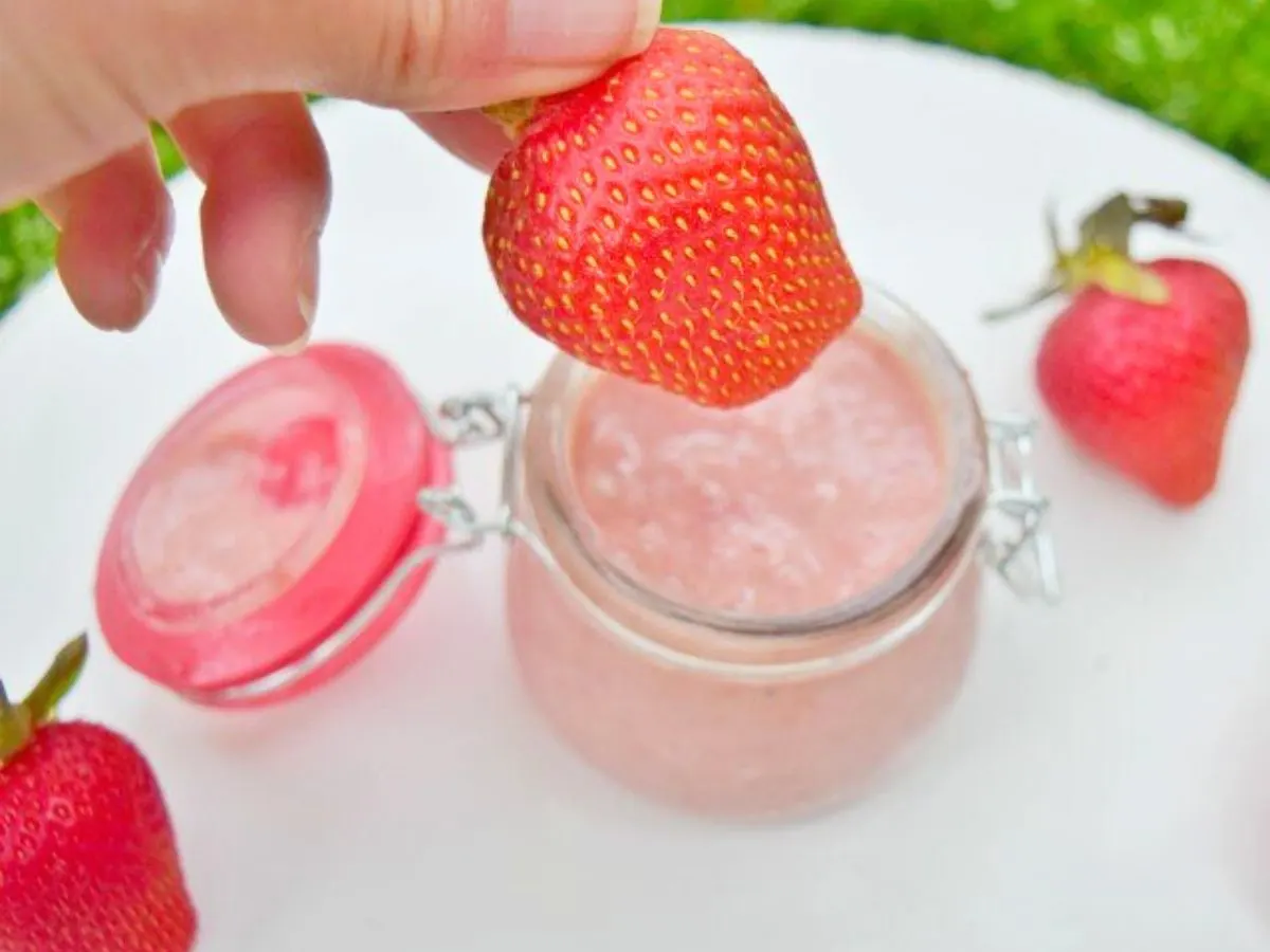 Image shows a hand holding a strawberry about to dip it into a small glass jar filled with strawberry curd.