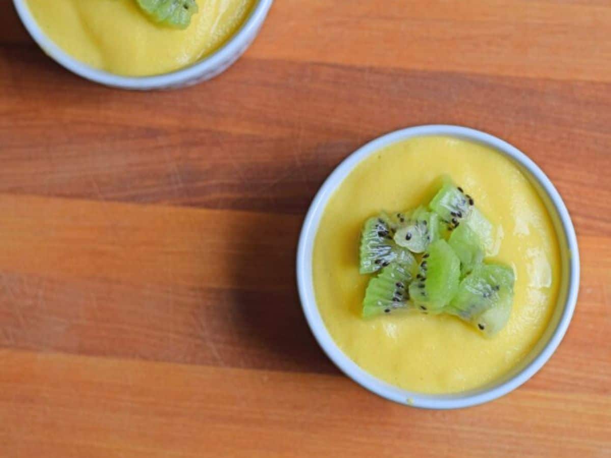 An overhead shot shows two ramekins with mango custard topped with chopped kiwi on a wooden cutting board.