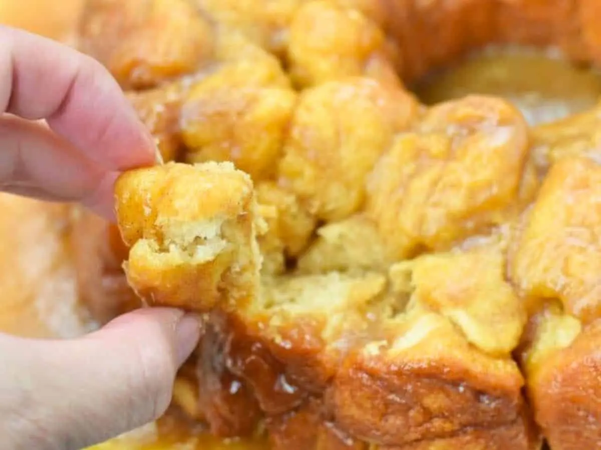 This photo shows a hand reaching out to monkey bread on a white cake platter and pulling off a chunk.