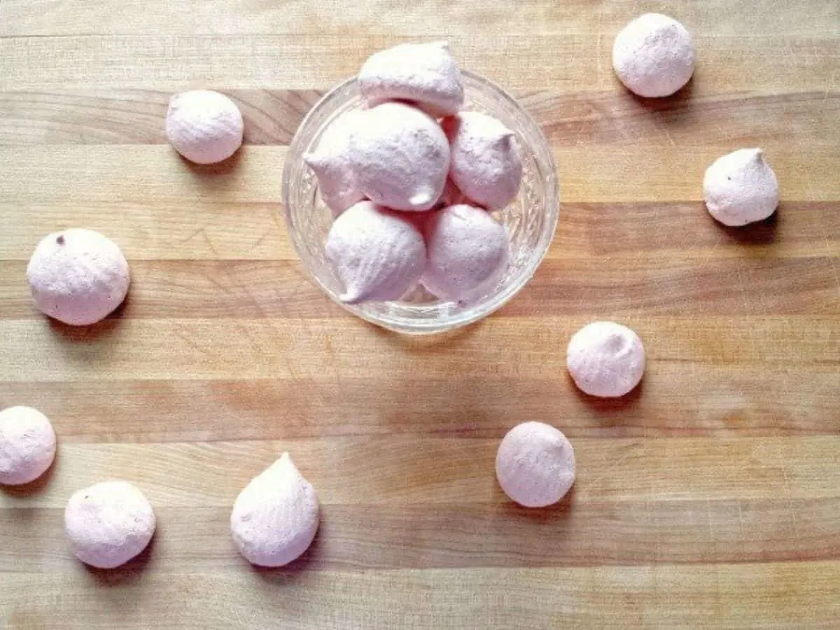 Image shows Raspberry Meringues in a glass parfait dish with more surrounding it on a wooden board.