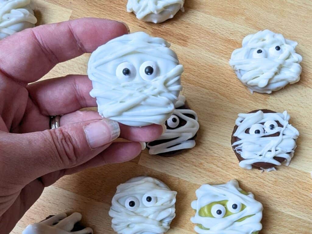 Image shows a Hand holding a mummy oreo cookie over others on a wooden board.