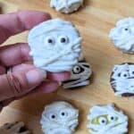 Image shows a Hand holding a mummy oreo cookie over others on a wooden board.