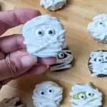 Image shows a Hand holding a mummy oreo cookie over others on a wooden board.