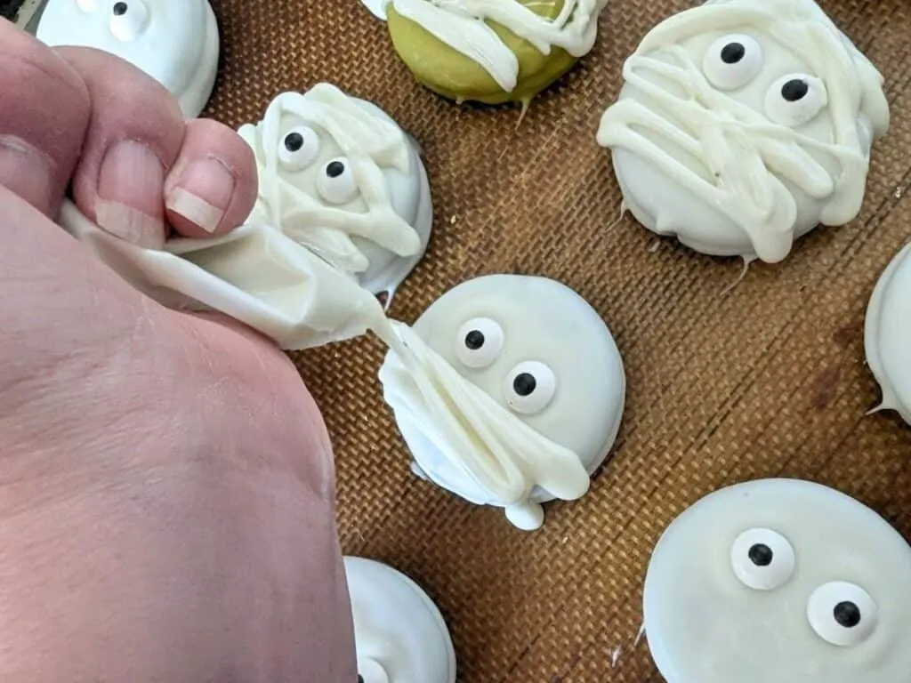 Image shows a hand Piping white chocolate on Halloween mummy oreos.