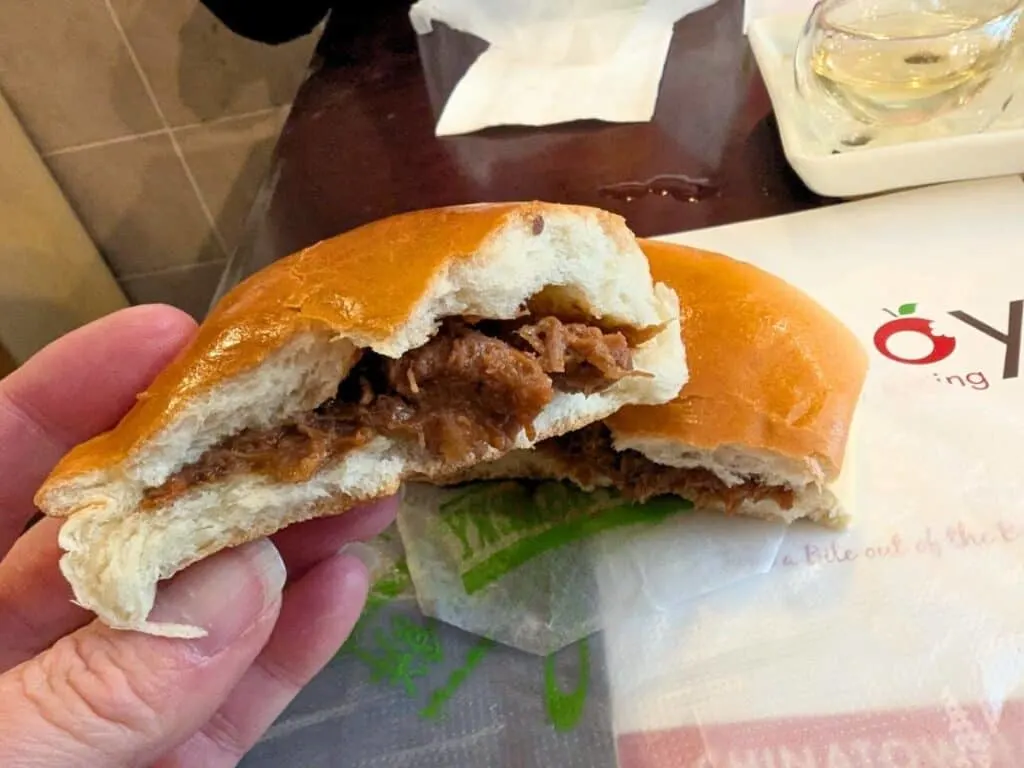 Image shows a hand holding half of a BBQ pork bao over a table with the other half.