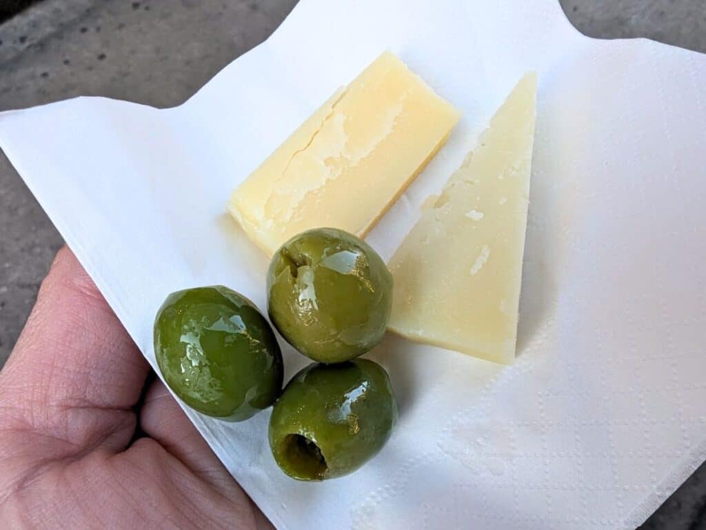 Image shows a hand holding a napkin containing two slices of cheese and three green olives.