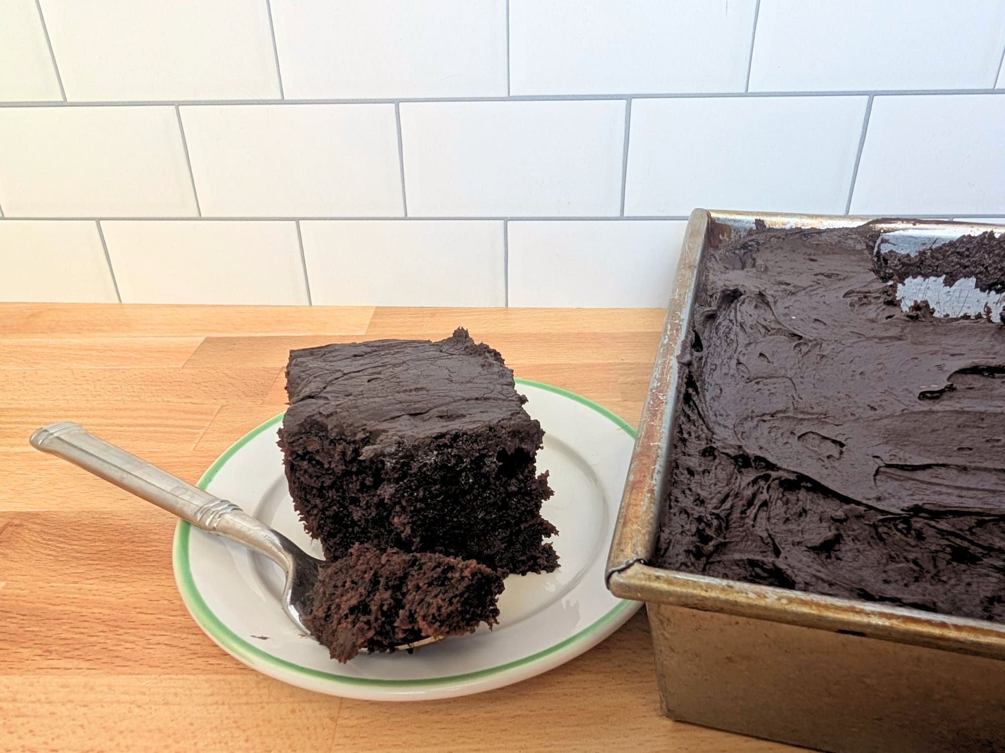 A slice of chocolate zucchini cake on a plate next to the rest of the cake in a 9x13 pan.