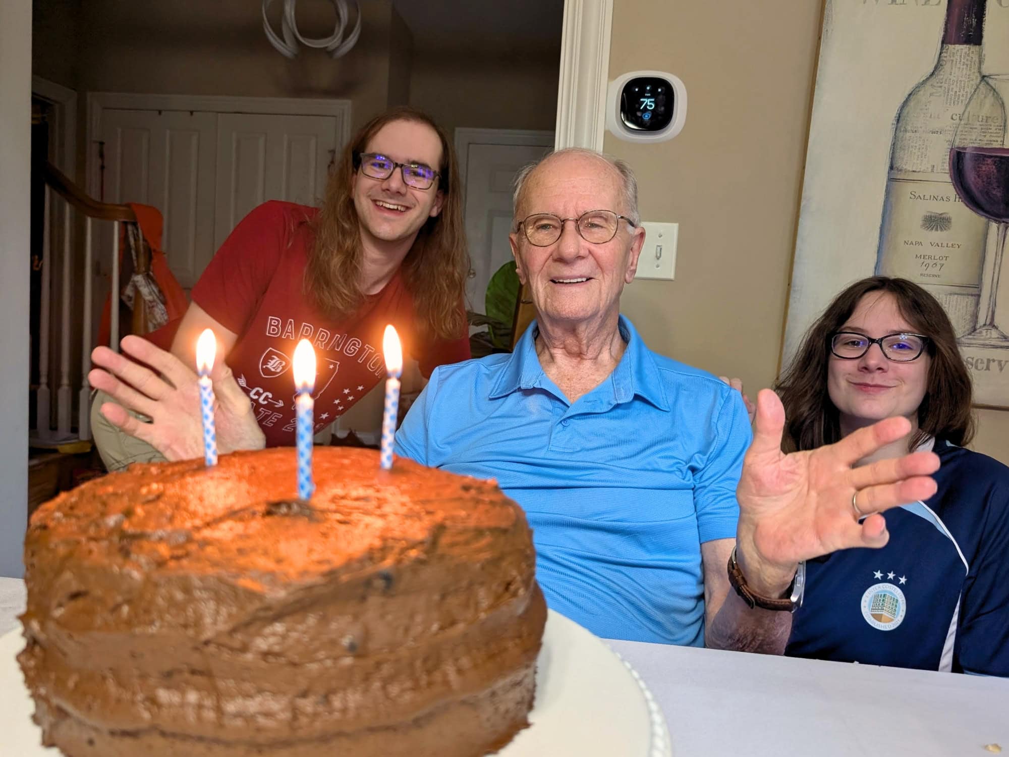An older man sits between two younger people behind a chocolate birthday cake with three lit candles.